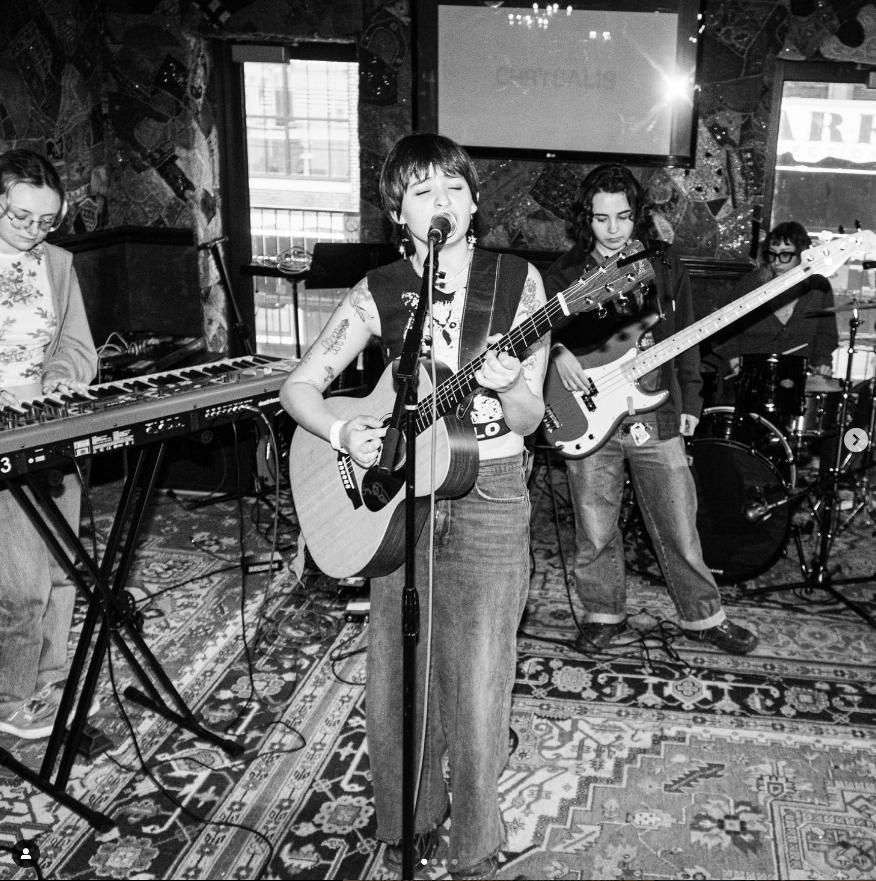 black and white photo of a thin light skinned person with short hair wering jeans and a sleeveless shirt playing guitar and singing with several other musicians acompanying them on various instruments