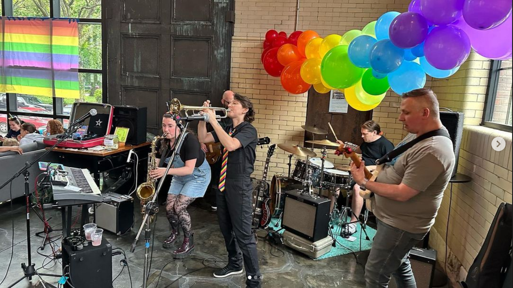 Doublestar performing with rainbow baloons above them and a rainbow flag in the window nearby. They consist of people playing saxophone, trumpet, drums, guitar, and bass. The saxophone player has light skin and dark hair and is wearing a black t-shirt with jean short overalls and patterned black and lace tights with boots. The trumpet player is wearing all black with a rainbow tie and is light skinned with neck-length hair. The bass player is bald with a beard and glasses. The guitar player has light skin, close-shaved hair and is wearing a grey t-shirt and jeans. The drummer has short hair that's longer on the top and is wearing a black t-shirt.