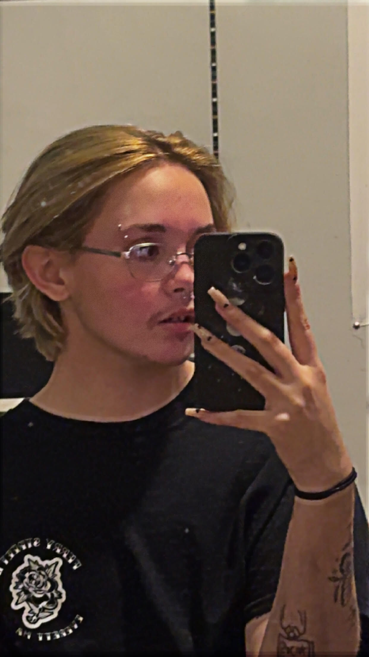 A white, shoulder length blonde, trans man with gold colored glasses with an eyebrow and septum piercings. He is wearing a black T-shirt. He is in front of a mirror, holding his phone, looking to the side.