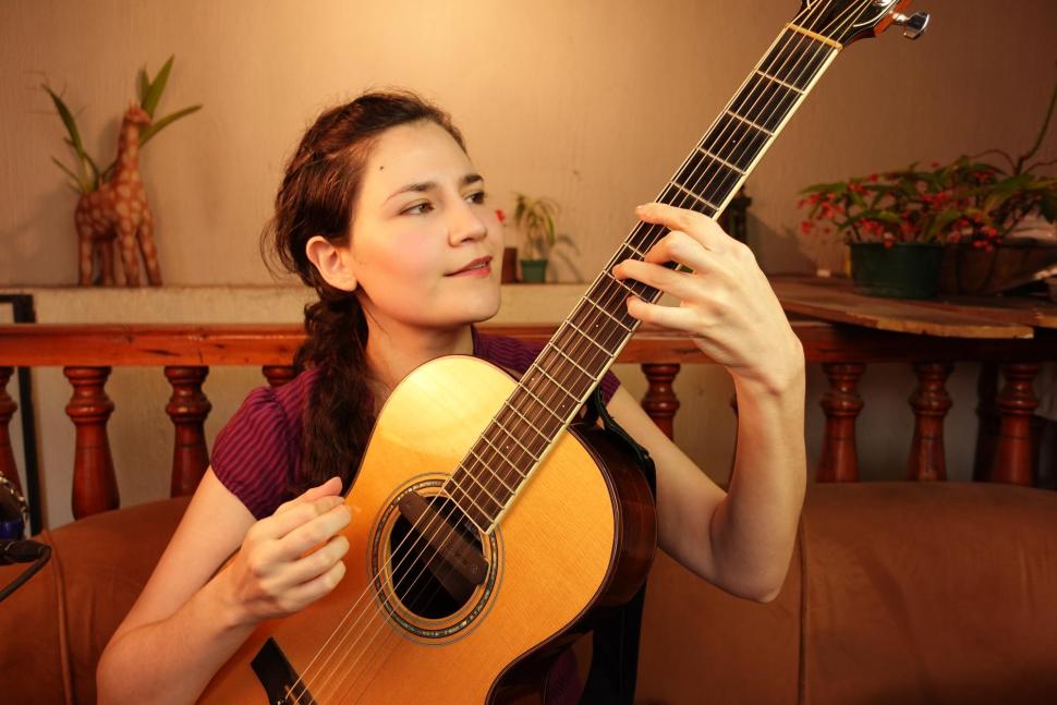 Light skinned brunette Guatemalan woman with hair pulled to the side smiling slightly and looking off camera while playing an acoustic guitar