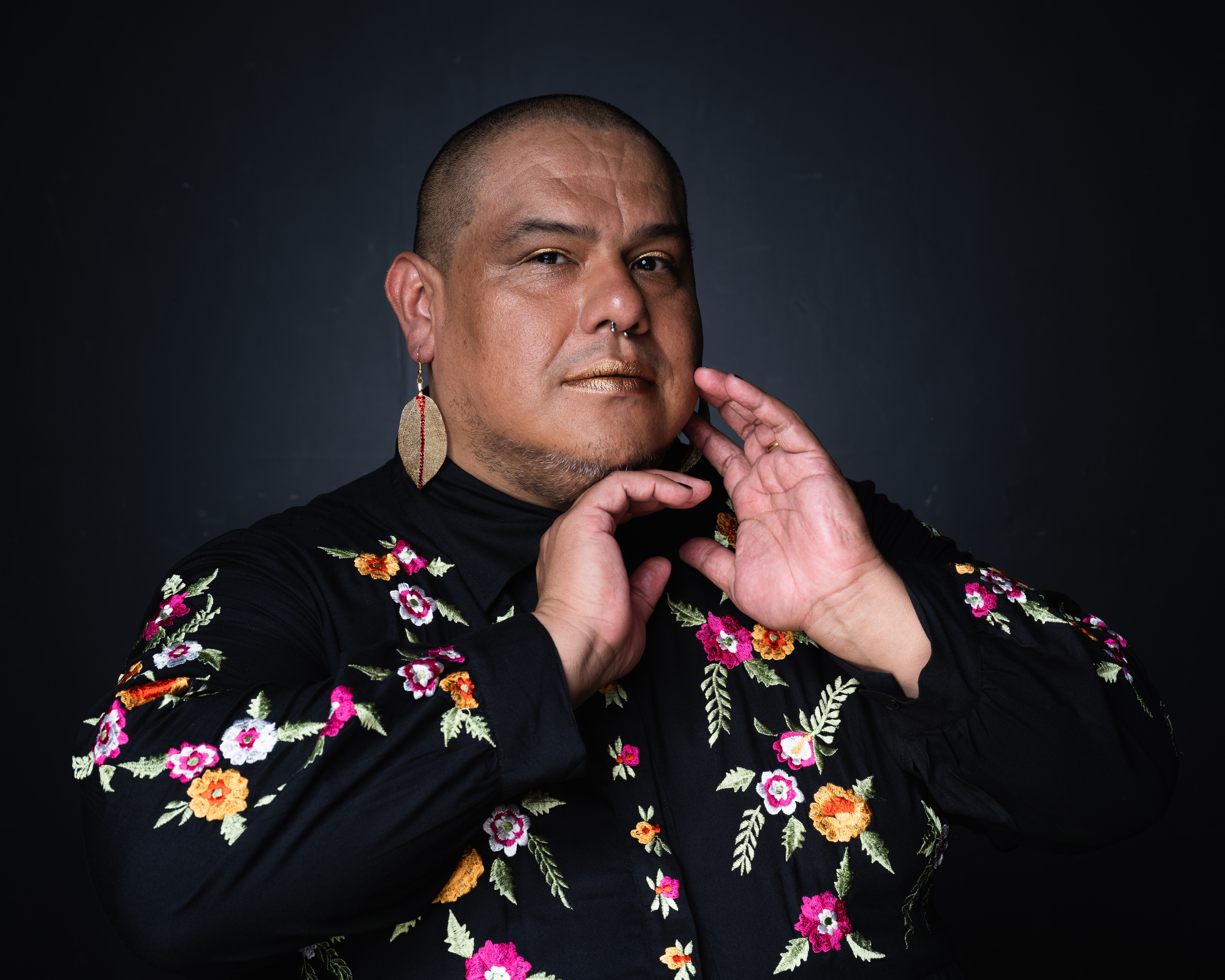 Medium shot of brown-skinned trans femme person with hands positioned in front of her face. Behind her is an accented dark gray background. She is wearing a long-sleeved black dress with multi-colored embroidered flowers and earrings in the shape of golden leaves.
