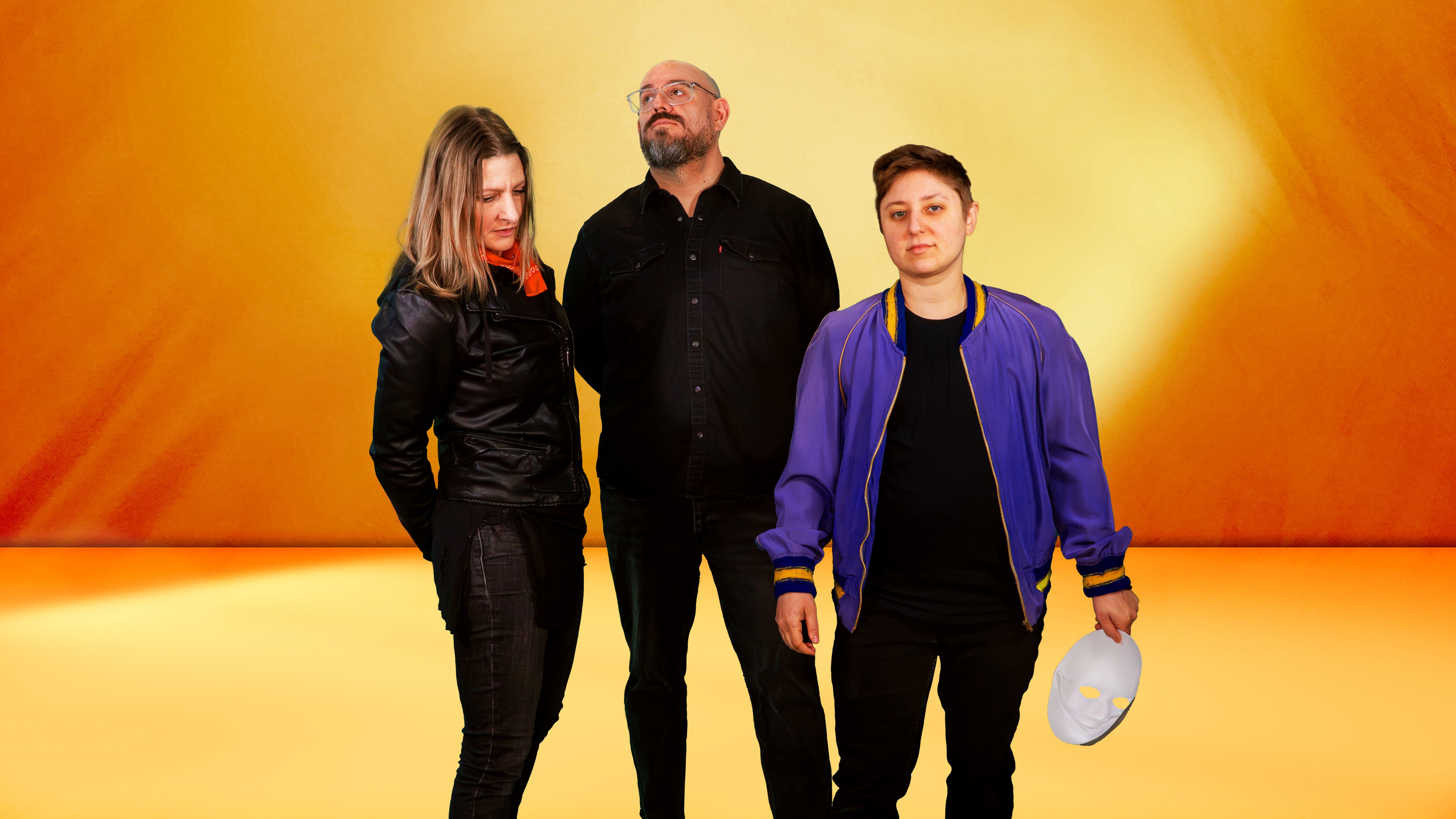 The three members of Happy Little Clouds standing in front of an orange backdrop, Jess Townsend on the left and Derek Bergstrom in the middle are looking off in different directions, but Jac Mestel on the right is looking directly into the camera lens.