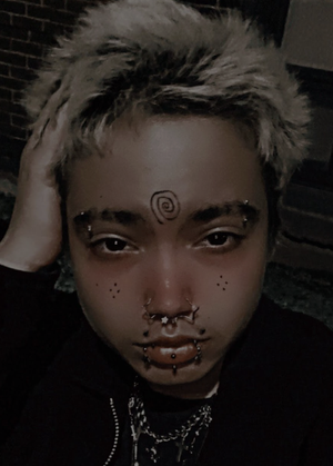 A very pierced trans man with light hair gazes directly into the camera.