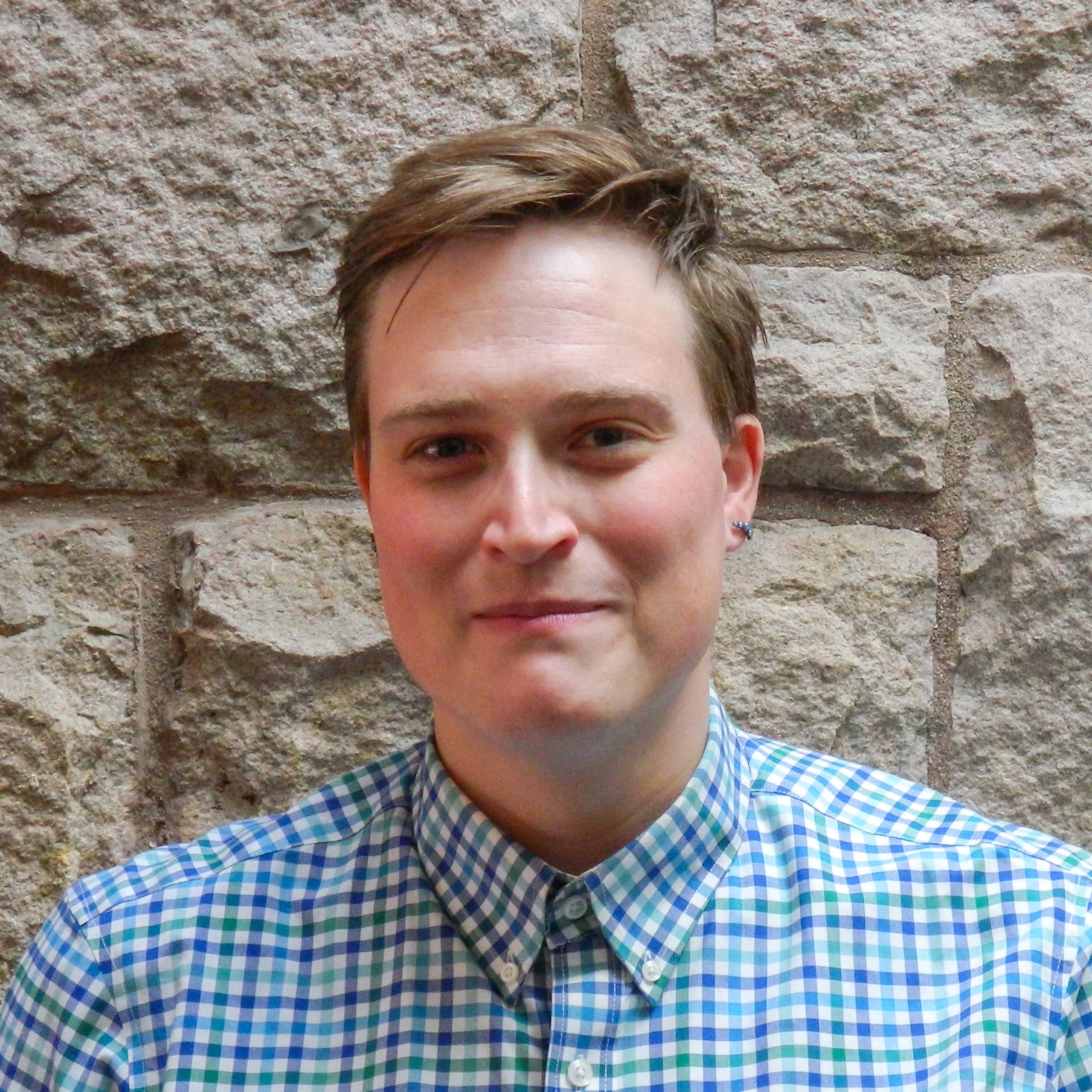 Photo of a butch white woman smiling at the camera. She is standing in front of a stone wall.
