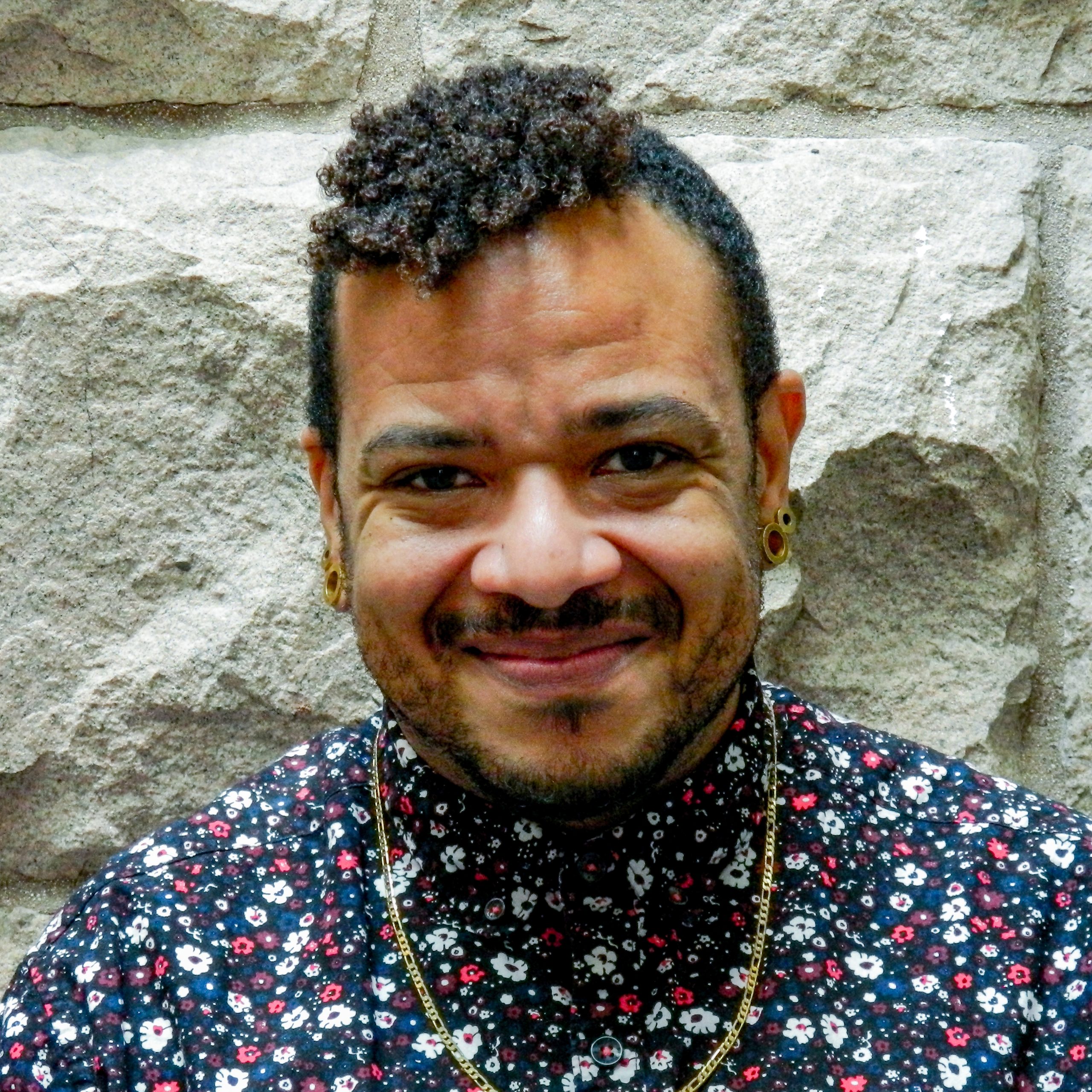 Photo of a Black man smiling at the camera. He is standing in front of a stone wall.