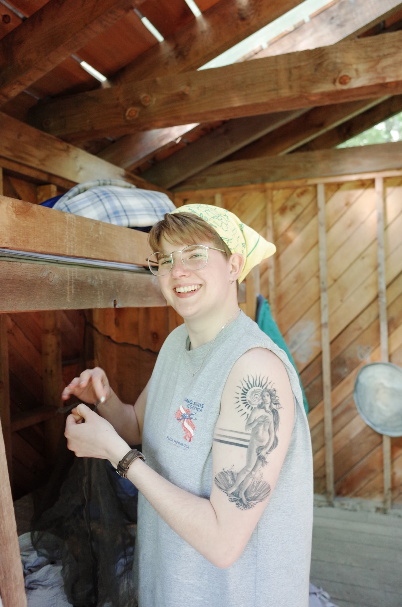 Photo of a white person with a light bandana and glasses wearing a grey sleevless shirt smiling at the camera. he has a large arm tatoo of a tastefully obscurred but mostly nude woman with long hair standing on some seashells with a stylized sun in the background.