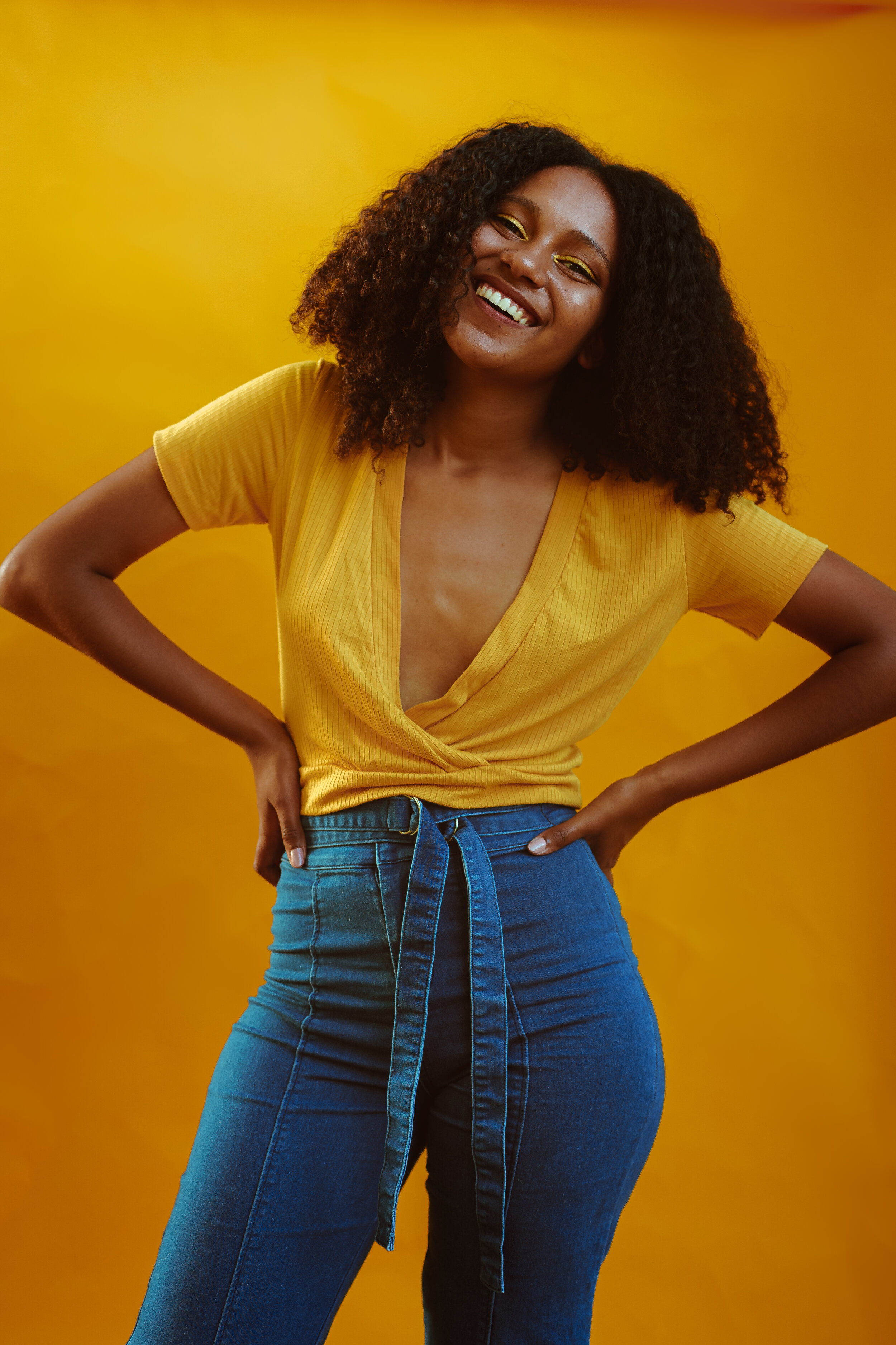 A Black Woman stands with her one hand on her hip, head tilted, smiling. The other hand is holding yellow flowers to the top of her head. She is wearing a yellow top and jeans and standing in front of a yellow background.