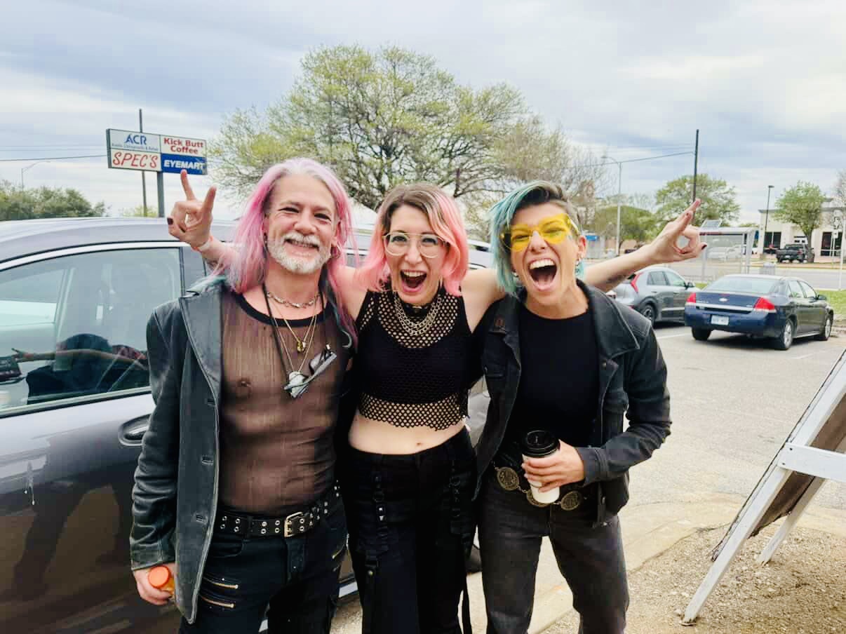 three people standing together in a parking lot. The one on the left has long pink and grey hair, grey facial hair, and is wearing as mesh shirt under a blue velvet-looking coat. The one in the middle has partially dyed pink hair and glasses with clear frames and is smiling with their arms around the other two people. They are wearing a black mesh cropped top with a black bra-like shirt and dark jeans. The last person has asymetrical light blue dyed hair, glasses with yellow lenses, and is wearing a dark jacket, black shirt, and faded black jeans. they are all smiling and look happy.