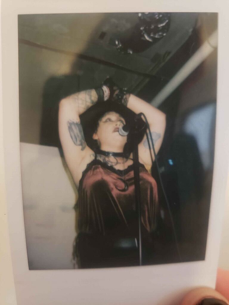 gothy feme with dark hair wearing a shimmering red/black dress and a collar with arms raised singing into a microphone