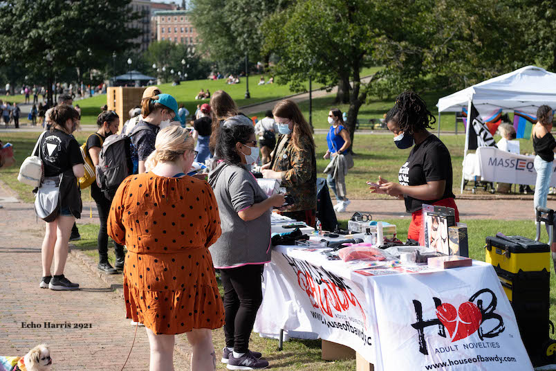 Dyke Fest attendees standing in front of tables. The table that can be seen by the camera is for House of Baudy, and adult novelties shop. Behind the table stands a black person with long hair braided and tied up at the back of the head. In front of the table stands a light skinned person wearing grey making a purchase. Waiting and looking at the table is a white person with blonde hair in an orange dress. A variety of lgbtq people are in the background.