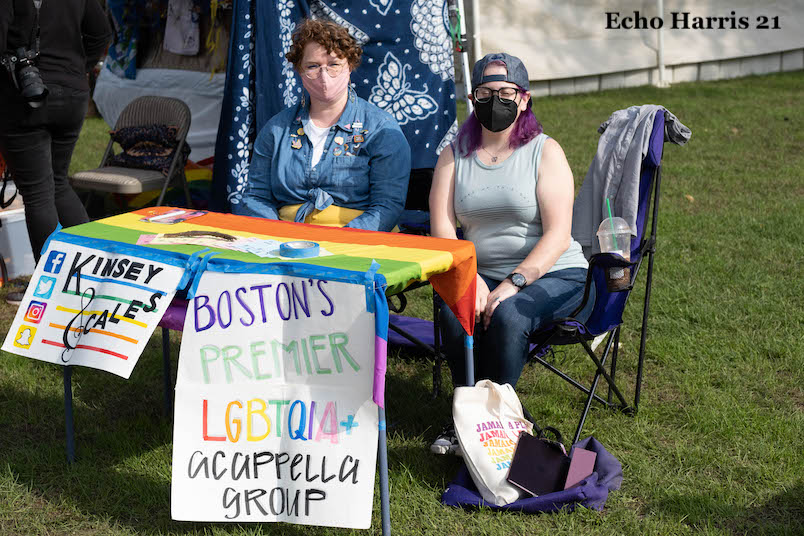 Photo of the Kinsey Scales table. Two white queer folks in masks sit behind a table with a rainbow flag. They are representing Kinsey Scales, Bostons premier LGBTQ acappella group