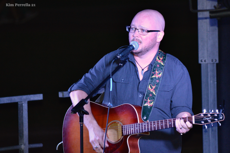 Photo of Kerr Griffin, a white bald transgender man with glasses and light stubble, playing guitar and singing into a microphone