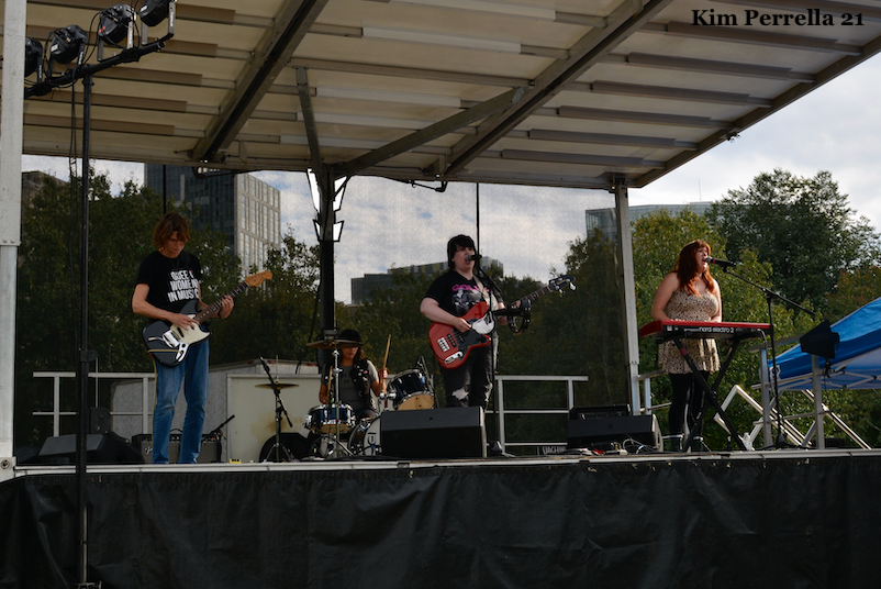 Photo of Thrust Club performing at Boston Dyke Fest. Thrust Club consists of four white women playing guitar, drums, bass, and keyboard. The lead guitarist is singing into the microphone.